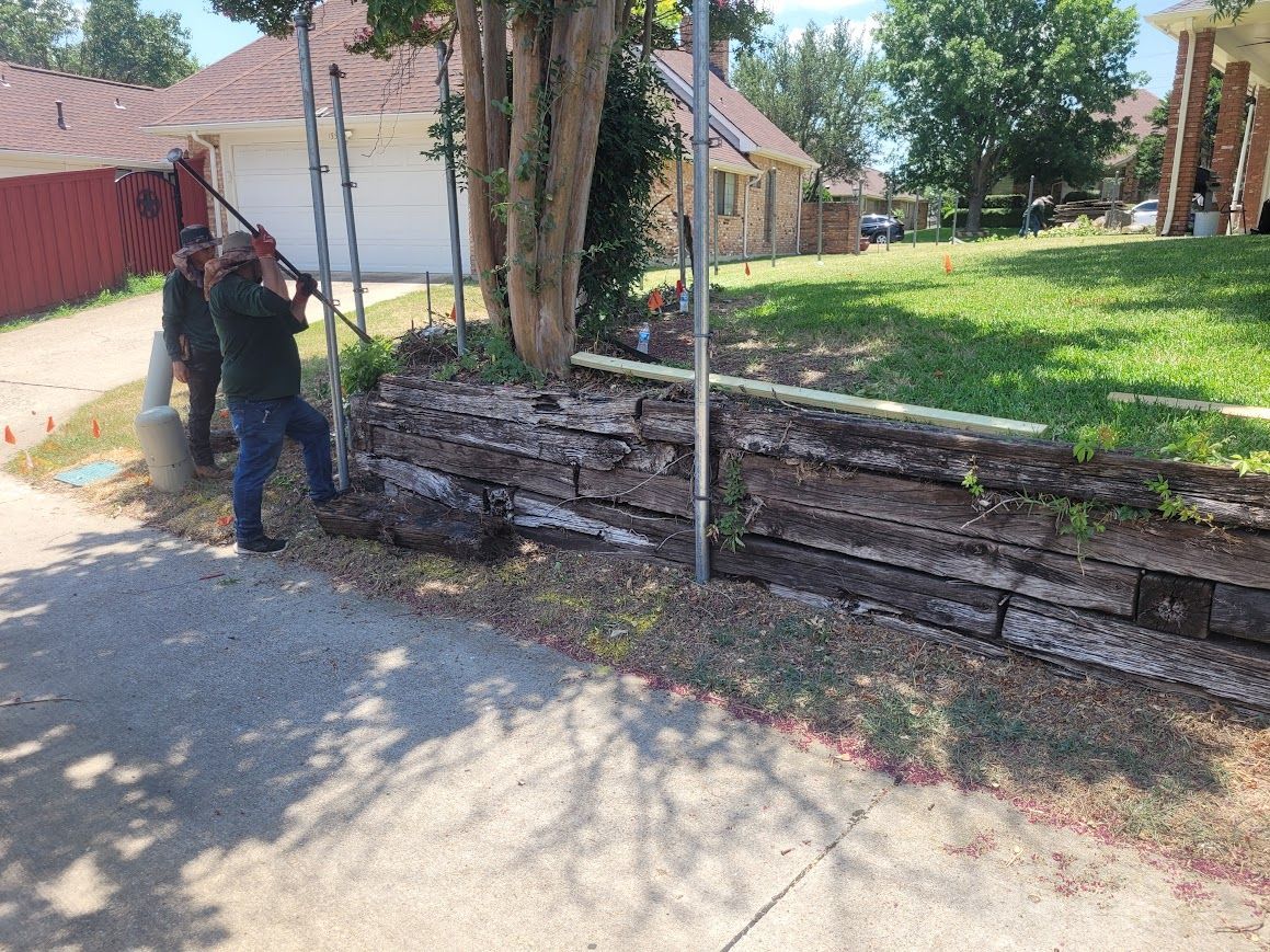 Person stands by a red fence and stone retaining wall near a tree-lined sidewalk.
