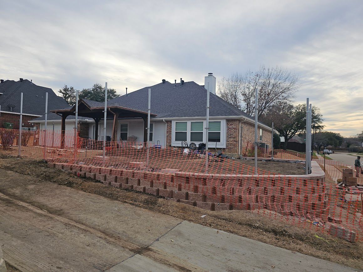 Single-story house with a gray roof and brick fence on a sloped street, under construction or renovation