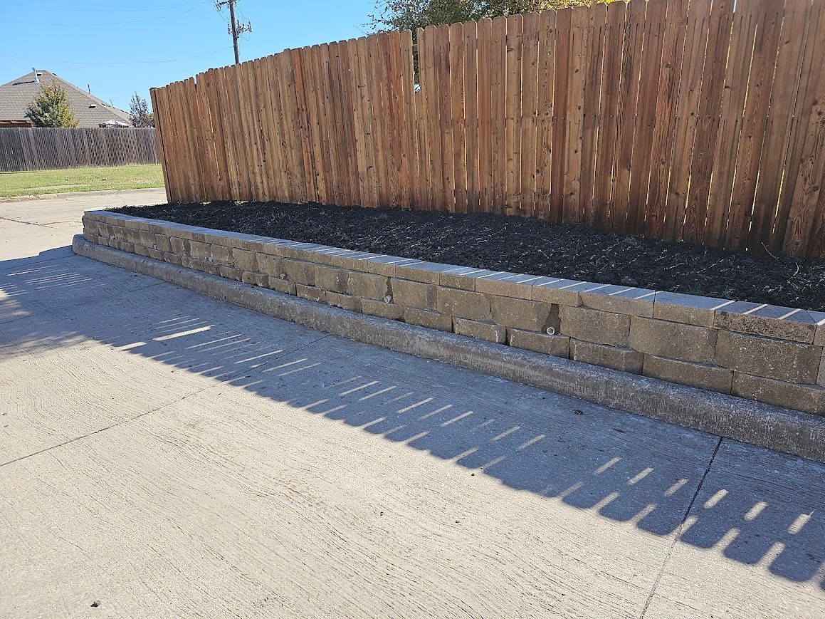 Stone retaining wall beside a wooden fence casting striped shadows on a paved roadside.
