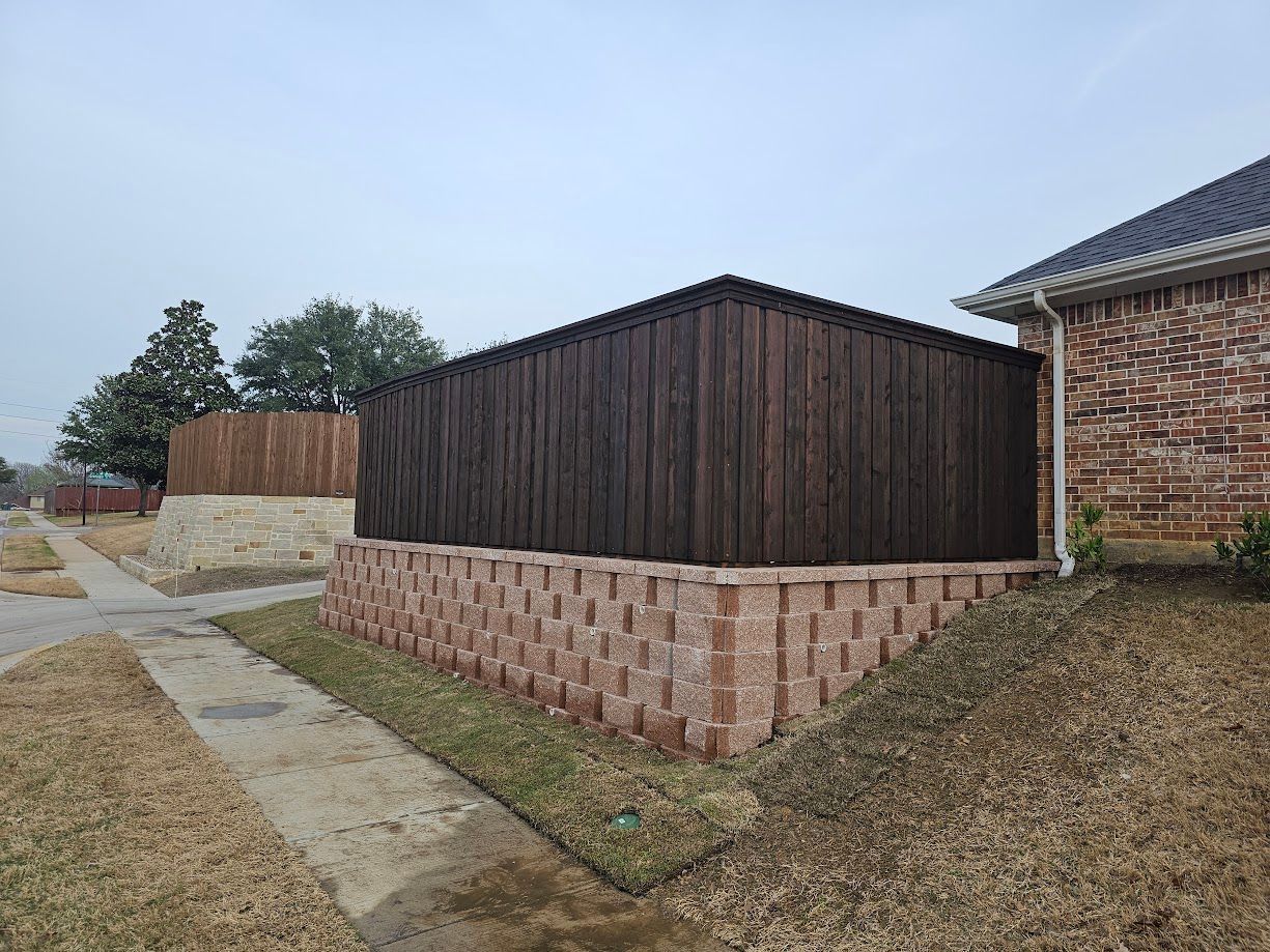 Outdoor view of a dark wooden shed on a brick base beside a concrete path and grassy yard