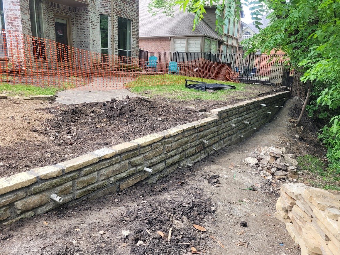 Stone retaining wall beside muddy creek behind a house with orange safety fencing and trees