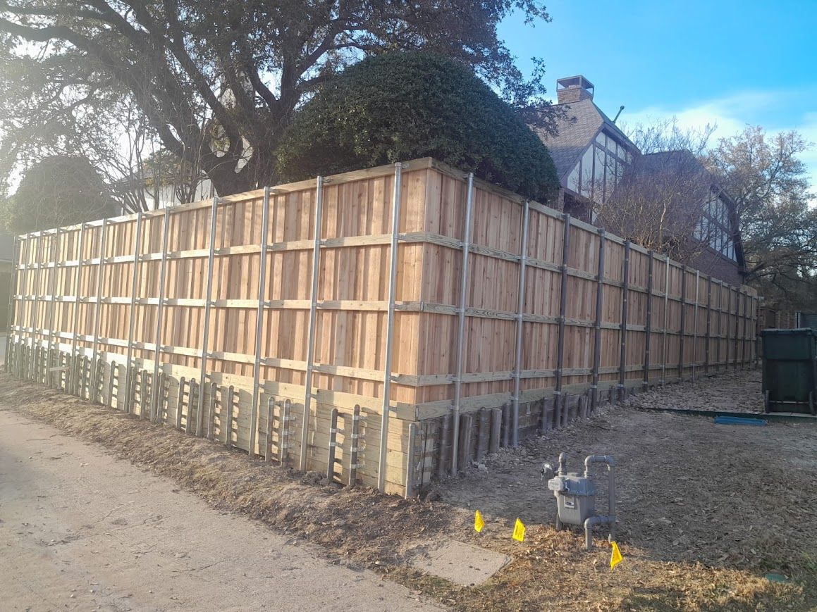 Wooden fenced enclosure beside a dirt road, with trees and a house in the background.