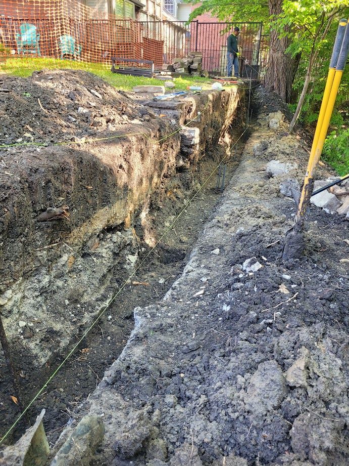Deep trench cut through muddy ground beside a brick fence, with a shovel and yellow post at right.