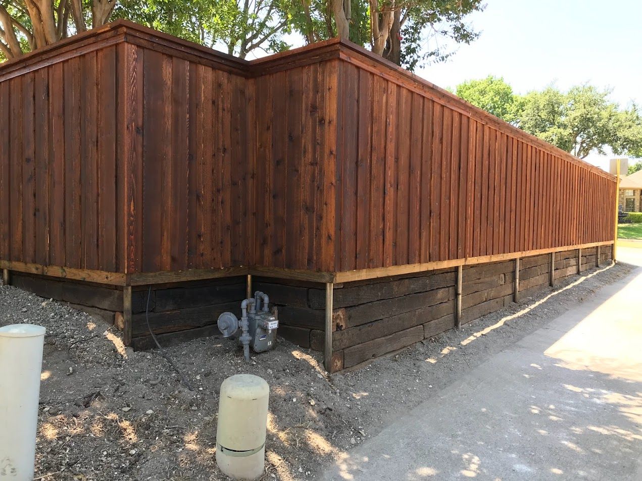 Wooden privacy fence along a sidewalk, with a black support frame and trees behind it