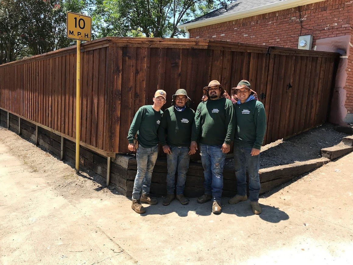 Four people in green uniforms standing beside a wooden fence at a corner, outdoors in bright sunlight.
