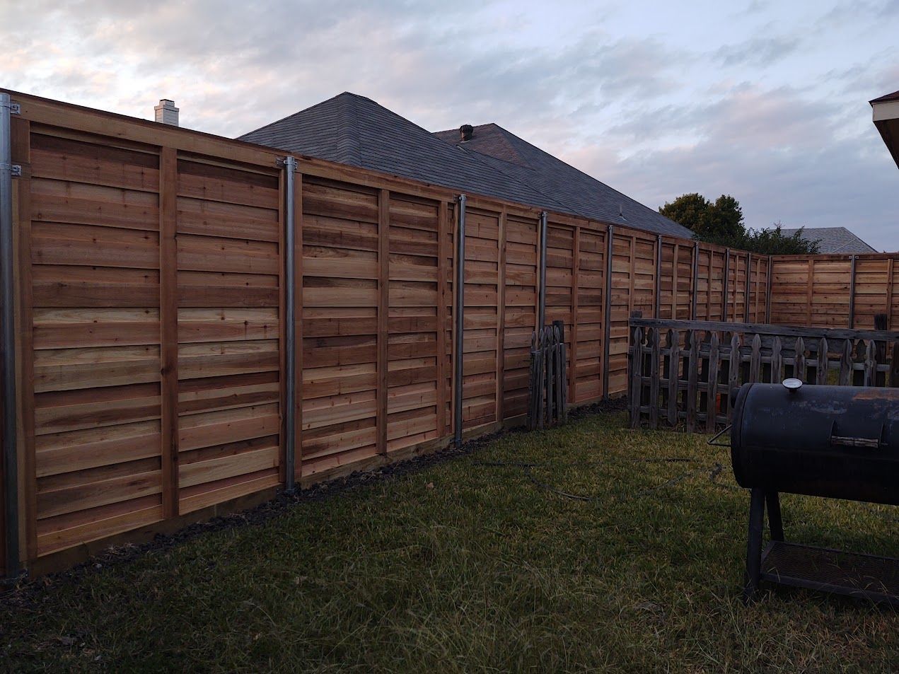 Wooden privacy fence along a grassy yard at dusk, with a black grill in the foreground.