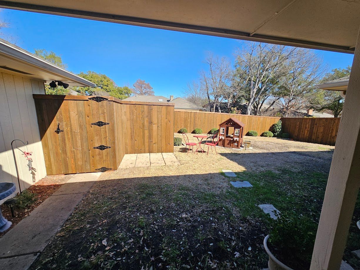 Backyard with wooden fence, gravel patio, small playhouse, and partly shaded lawn under blue sky
