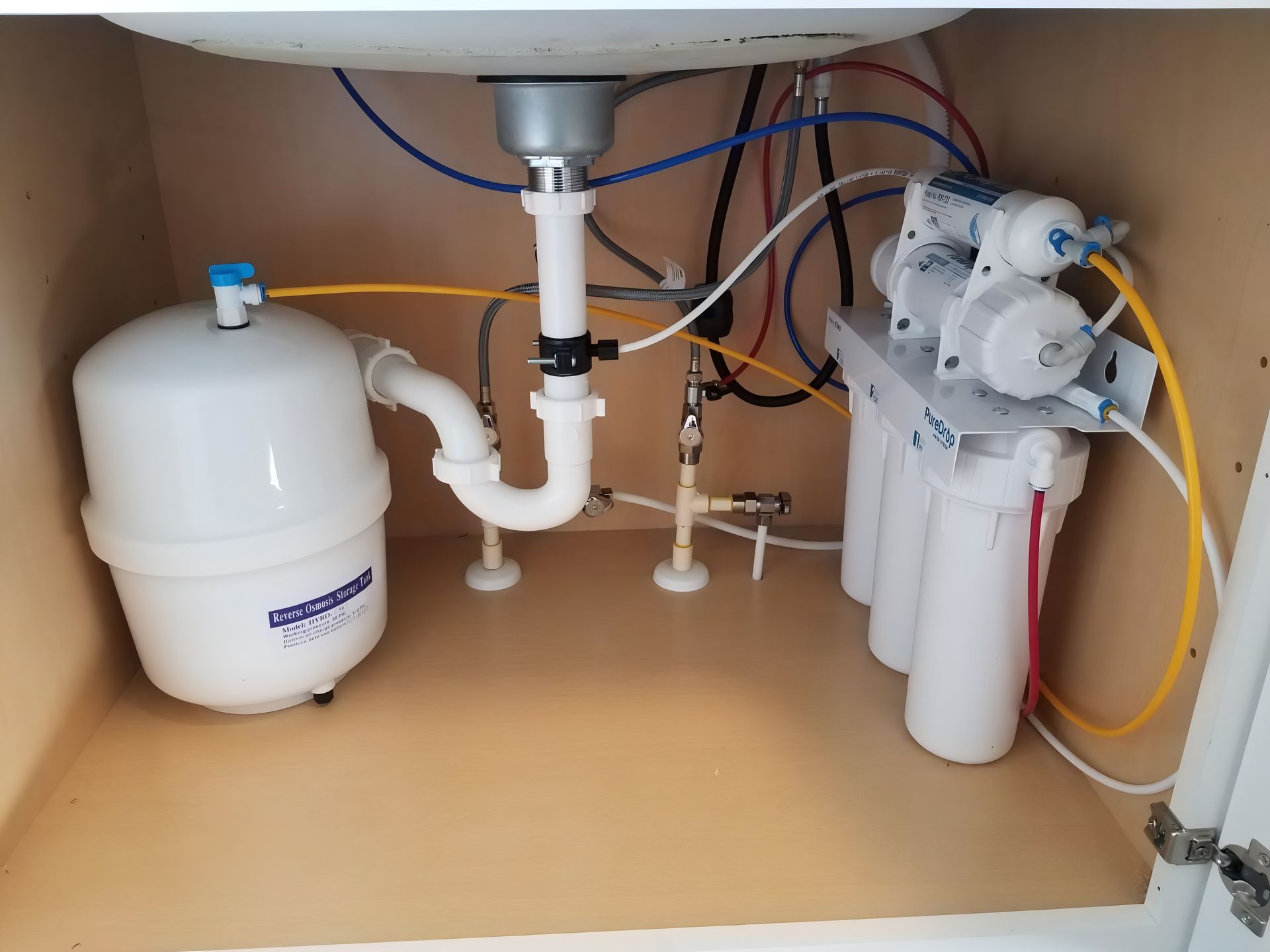 Inside a kitchen cabinet, an under-sink reverse osmosis water filtration system with a white storage tank and filters.
