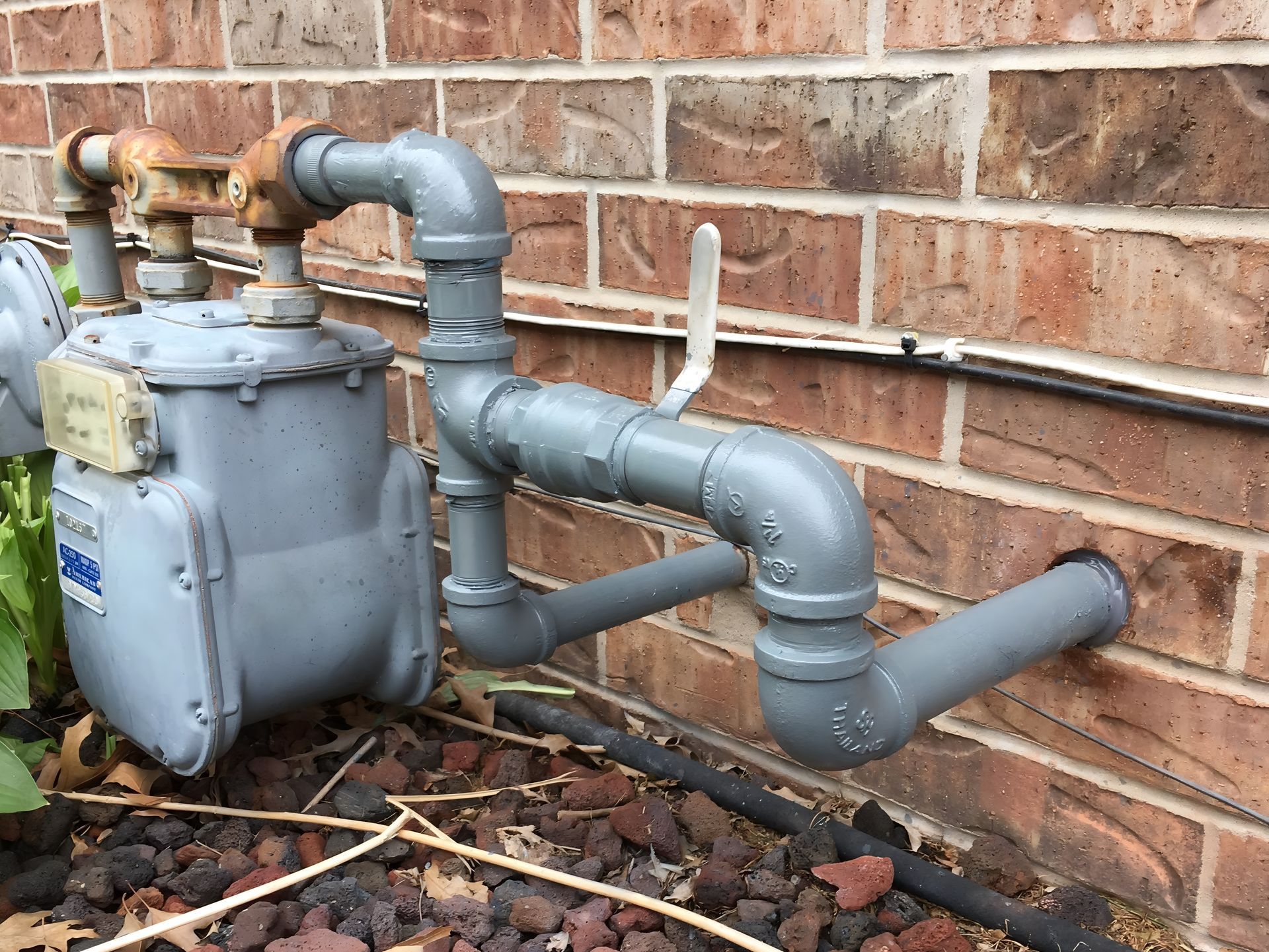 Gas meter and piping attached to a brick wall. Gray metal pipes, a white valve, and a black pipe run horizontally.
