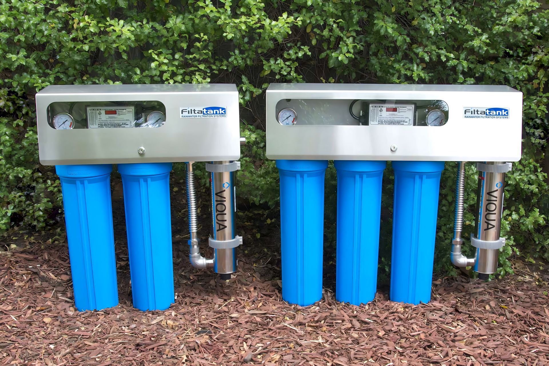 Two blue multi-filter water treatment units outdoors on mulch against greenery