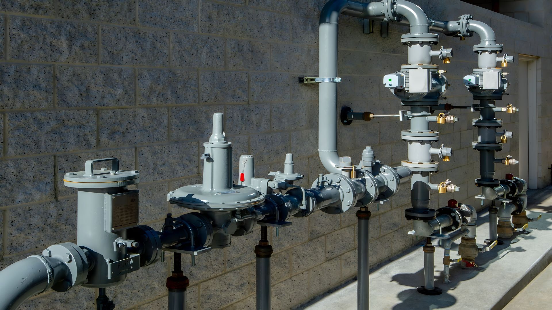Industrial piping and valves against a cinderblock wall, with a horizontal pipeline in the foreground
