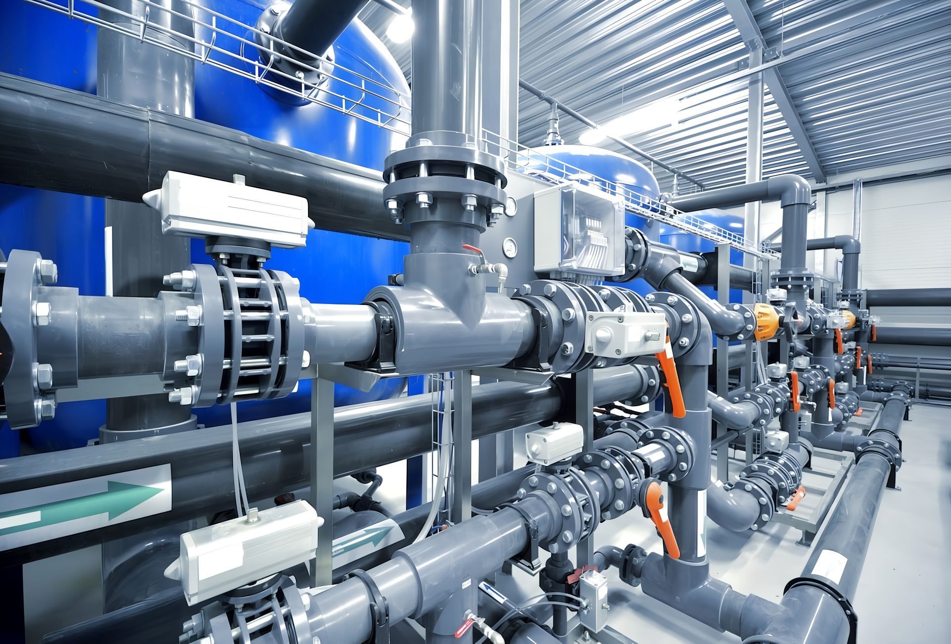 Piping system in a water treatment plant, featuring blue tanks, gray pipes, and valves, viewed indoors.