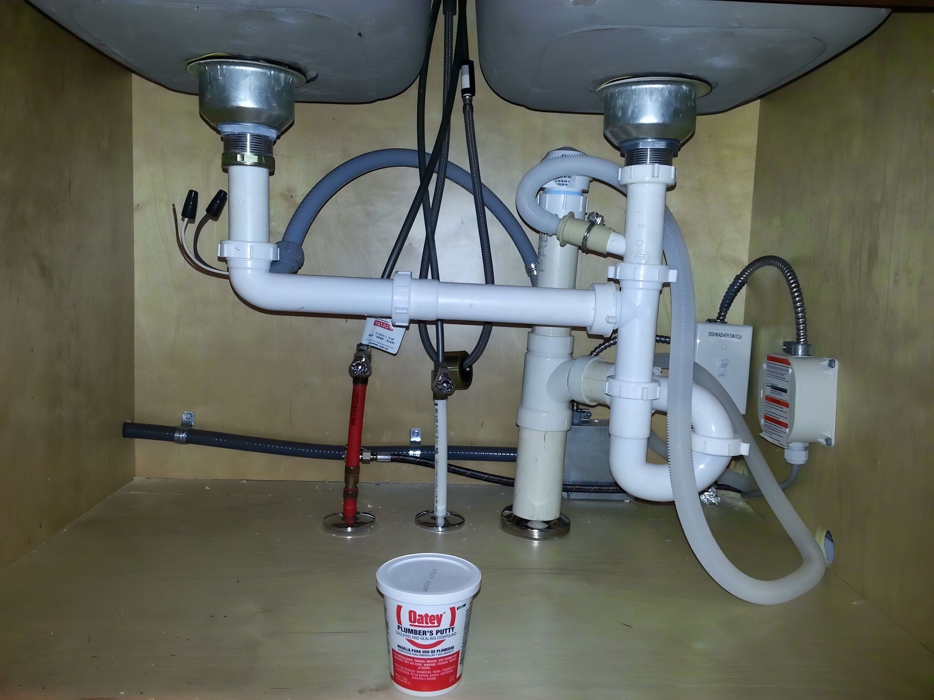 Under-sink plumbing with white pipes, valves, and a bucket placed below a double sink.