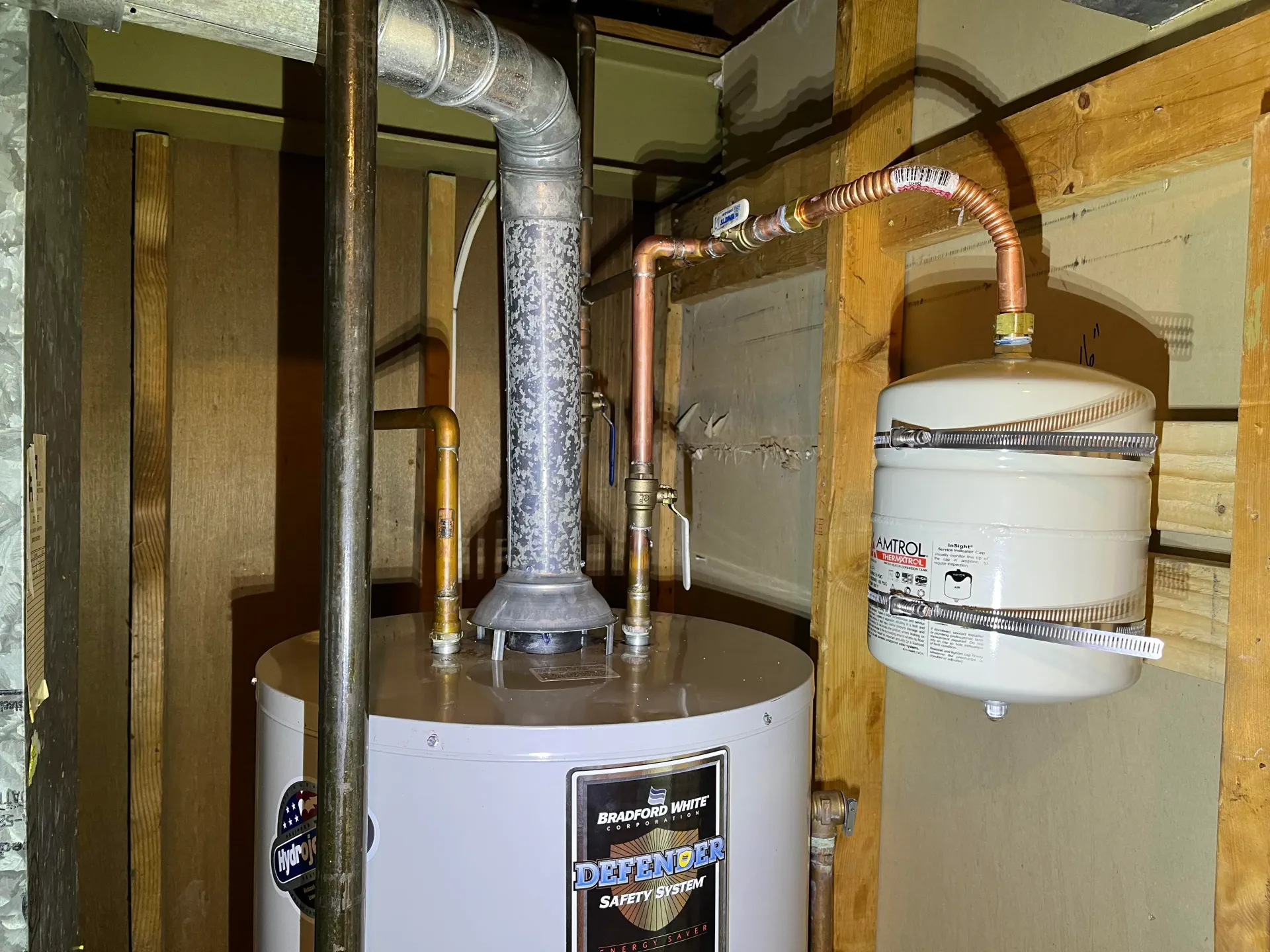 A water heater in a utility room with copper piping and a white expansion tank mounted on the wooden frame.