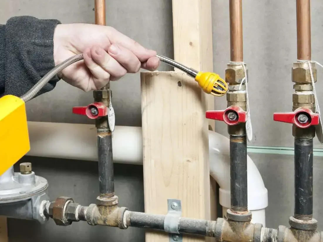 Person using a leak detector wand on copper pipes with red handled valves in a utility room.
