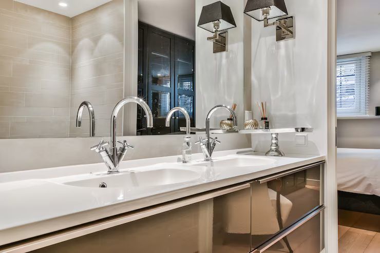 Bathroom with double sinks, chrome faucets, and a large mirror. Two lamps flank the mirror, and a bedroom is visible in the background.