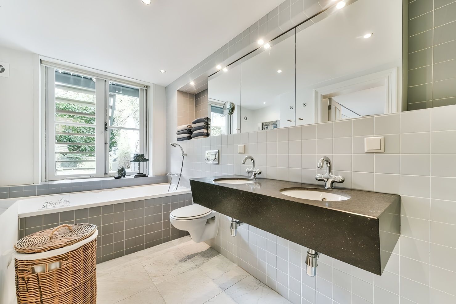 Modern bathroom with gray tile, a double sink, and a large mirror. A tub and window are visible on the left.
