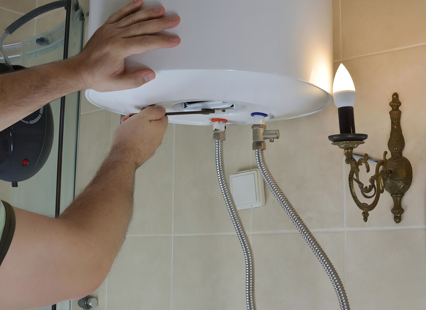 A person installing a water heater in a bathroom, using tools. The heater is white with silver hoses and brass wall sconce.