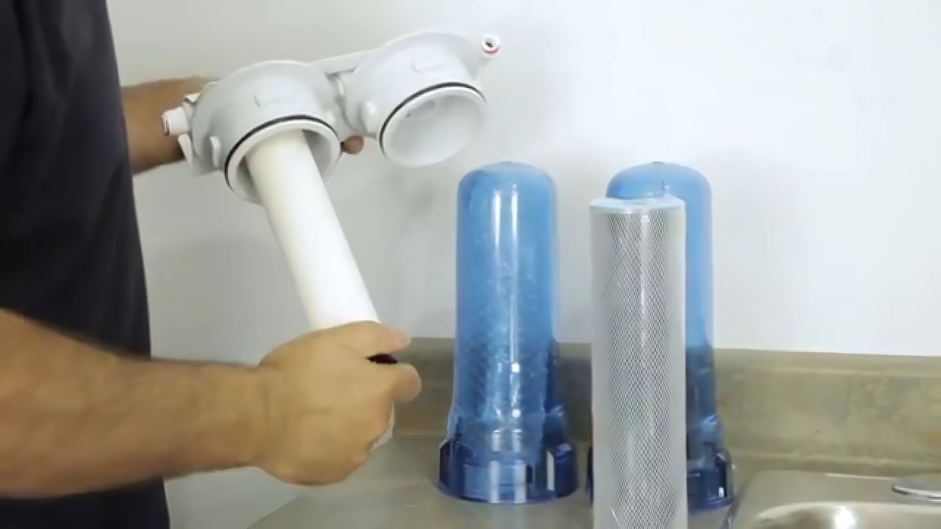 A person holding a water filter housing, preparing to install a new filter. The housing is white, and the replacement filters