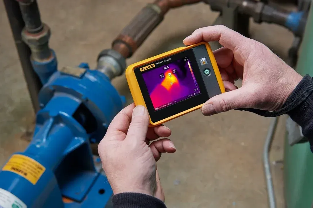 A person uses a handheld thermal camera to inspect machinery, showing a heat map on the screen.