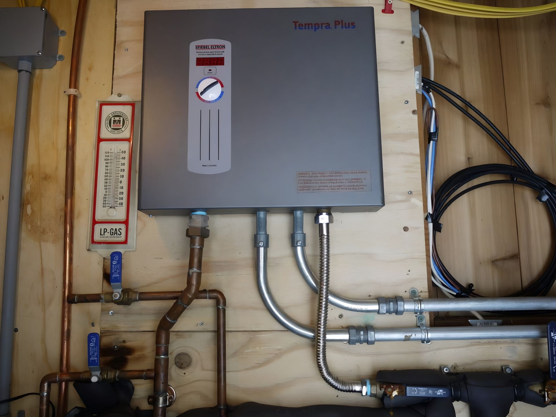 A gray tankless water heater with attached pipes and controls mounted on a wooden wall.