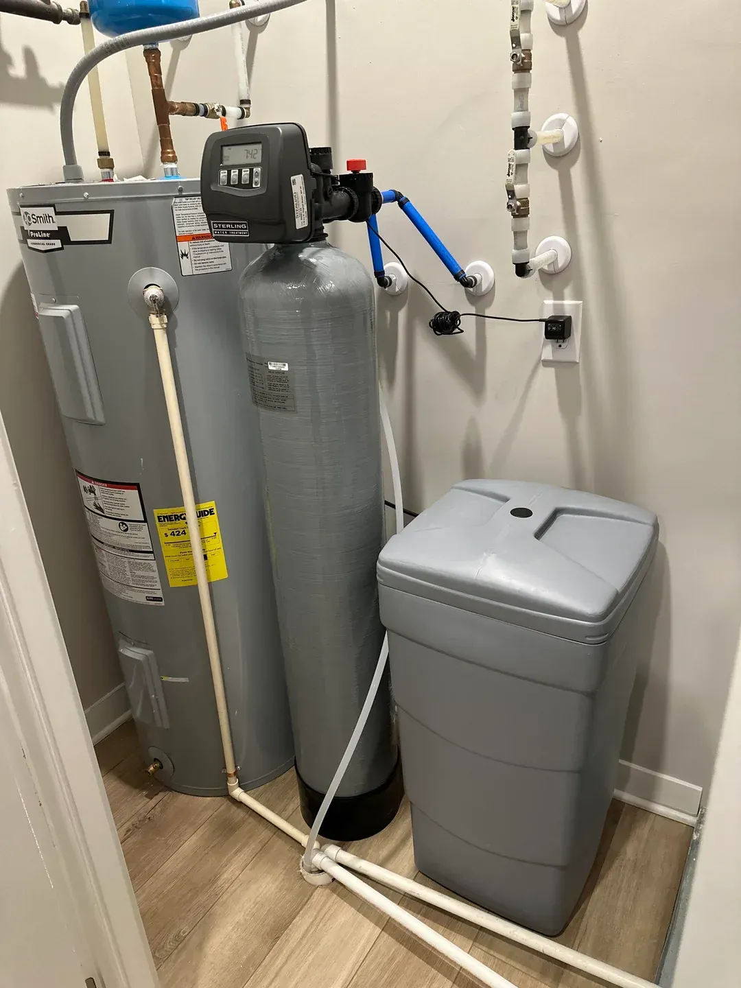 Laundry room with water softener and utility tanks beside a gray trash bin and exposed pipes.