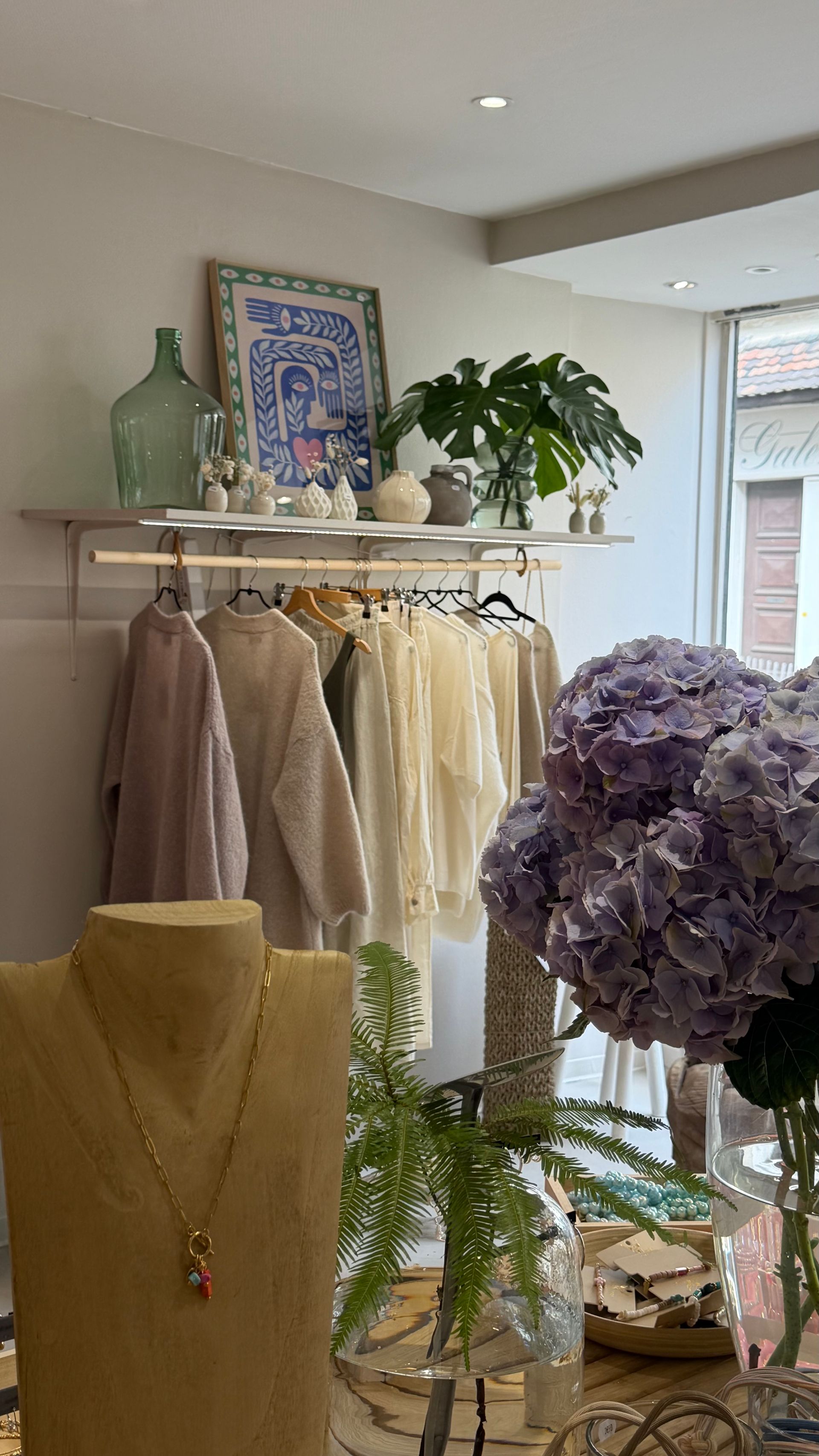 Présentoir de vêtements de boutique avec robes pâles, buste à bijoux, plantes et hortensias violets près d'une fenêtre