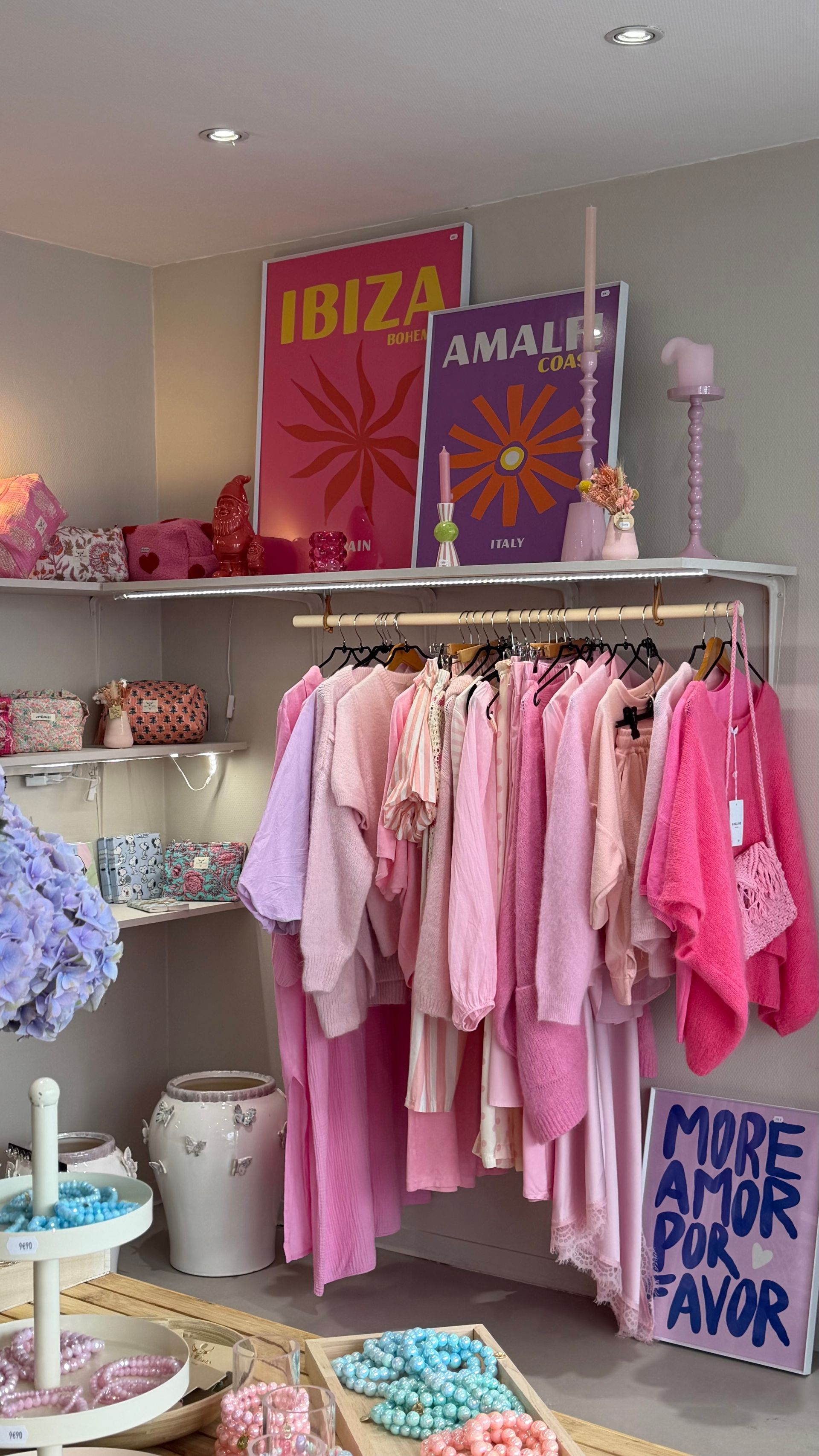 Un portant à vêtements rose dans une boutique avec des enseignes colorées « Ibiza Amarte » et « More Amor Por Favor ».