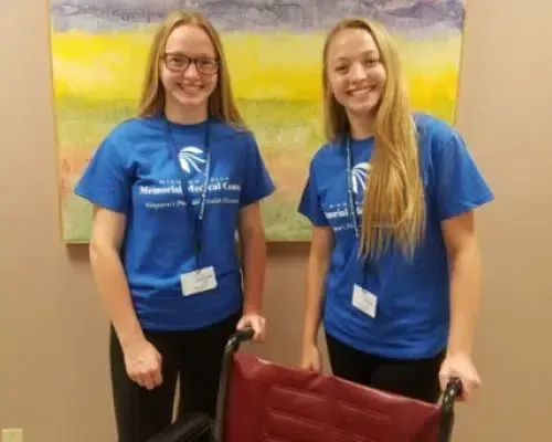 Two smiling people in blue T-shirts stand indoors in front of a colorful painting.