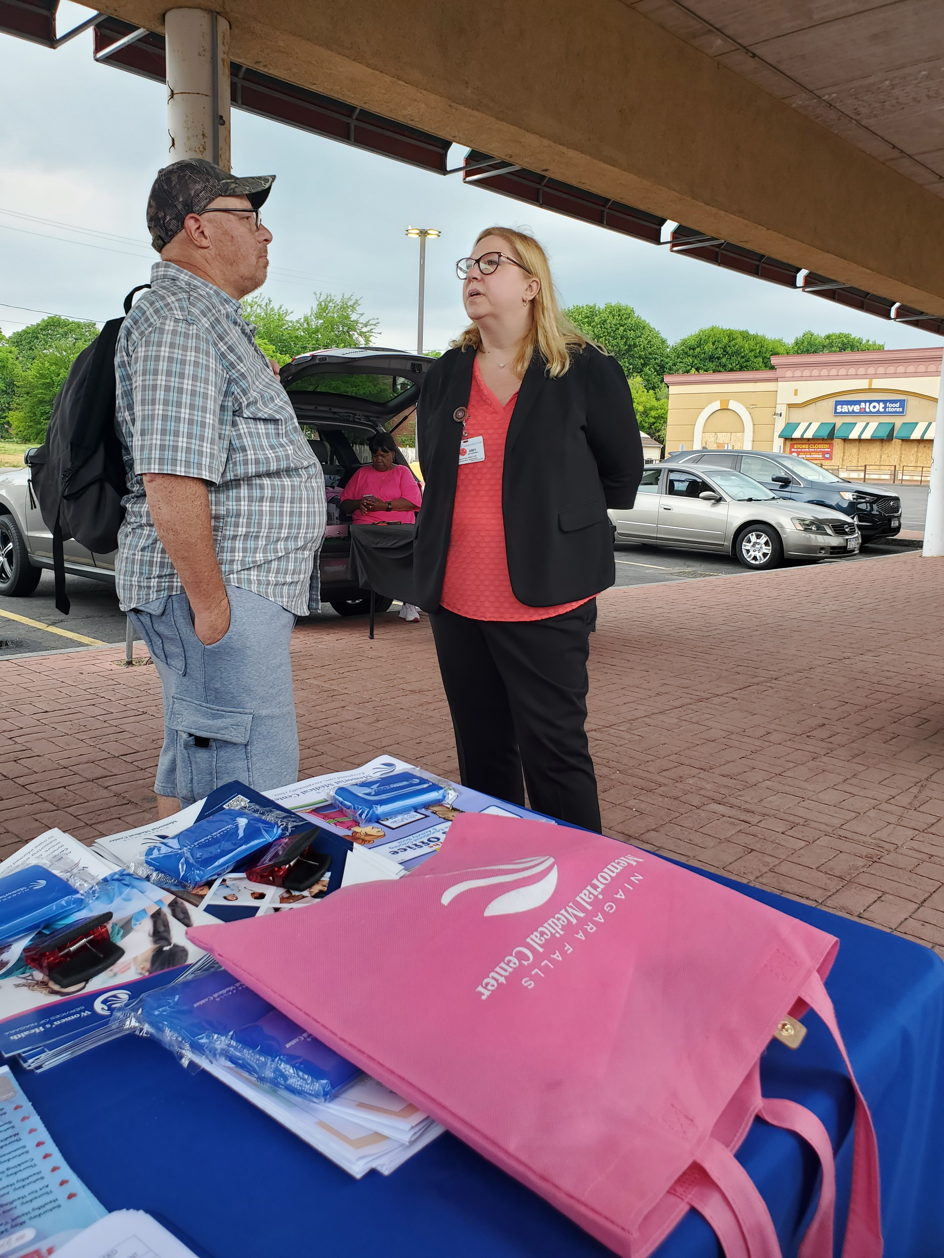Two people talk under a canopy beside a table of colorful bags at an outdoor market.