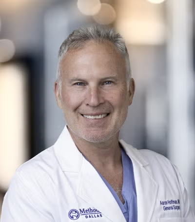Smiling medical professional in white coat and blue scrubs, standing in a hospital hallway.