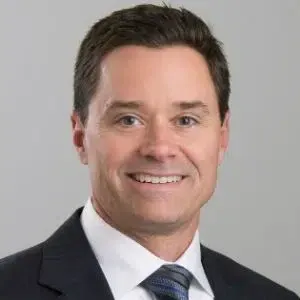 Smiling man in a dark suit and blue tie against a light gray background