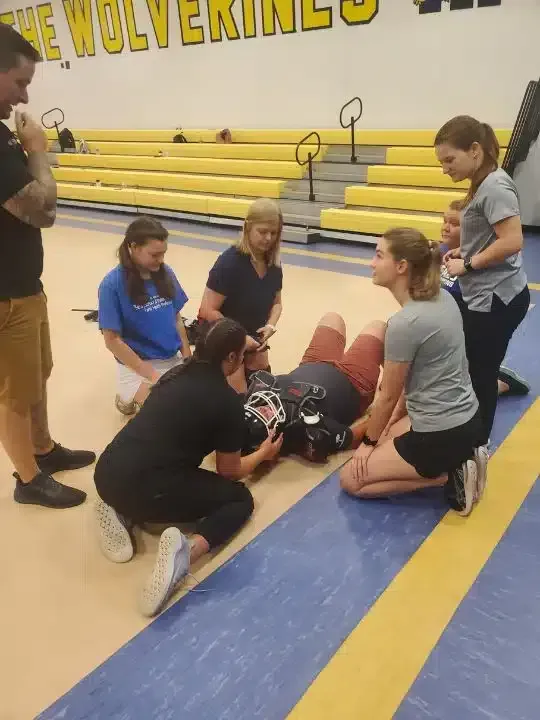Students and coach tending to an injured player on a gym floor, with bleachers in the background