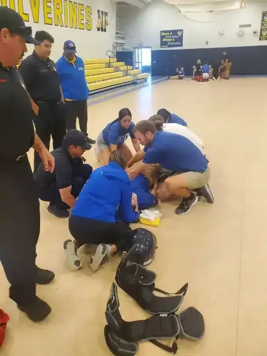 Students in blue shirts and pads practice on a gym floor while a coach watches nearby
