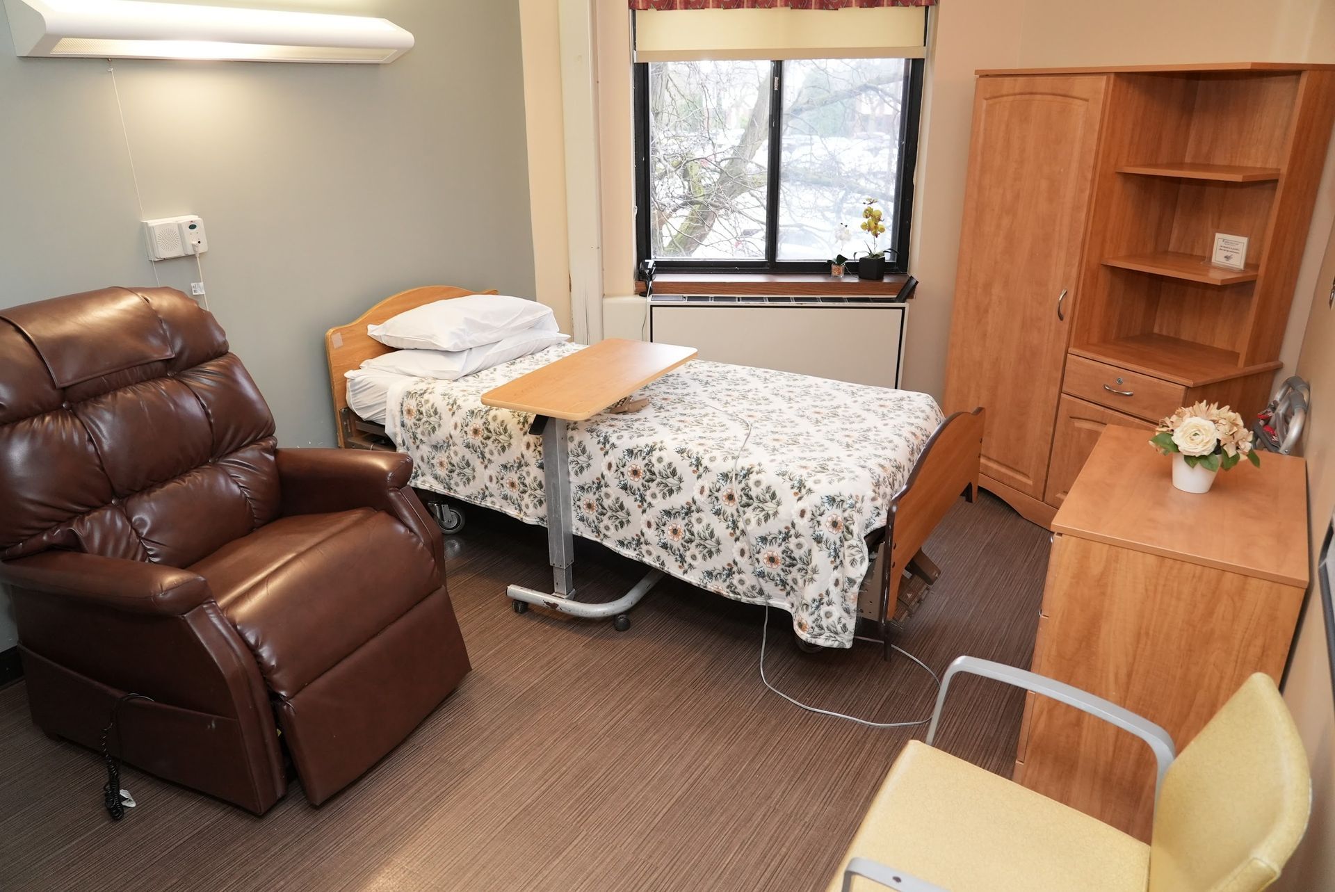 Small medical exam room with a bed, brown recliner, wooden cabinet, and window.