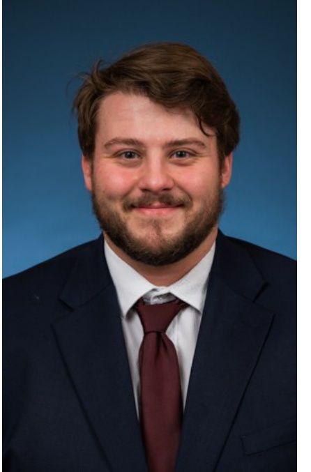Studio portrait of a smiling man in a dark suit and maroon tie against a blue background