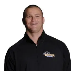 Man in a black zip-up jacket with a logo, standing against a white background and smiling.
