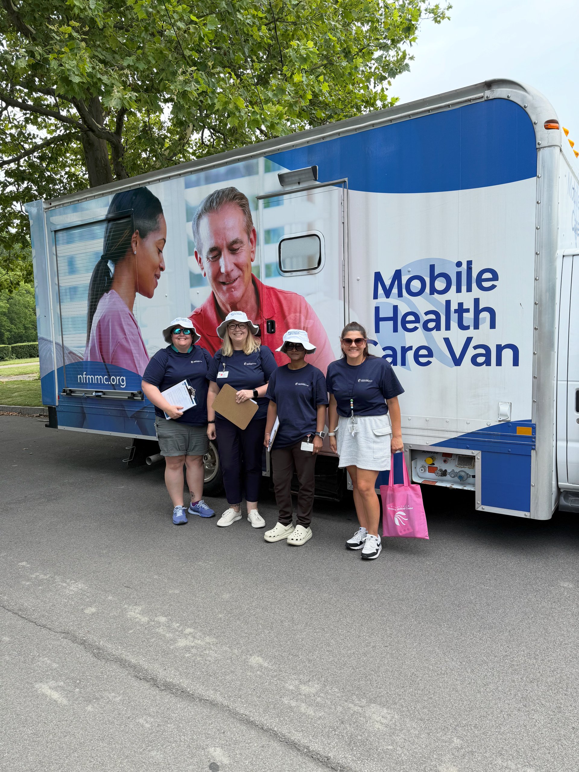 Four people pose beside a blue-and-white Mobile Health Van on a tree-lined street.
