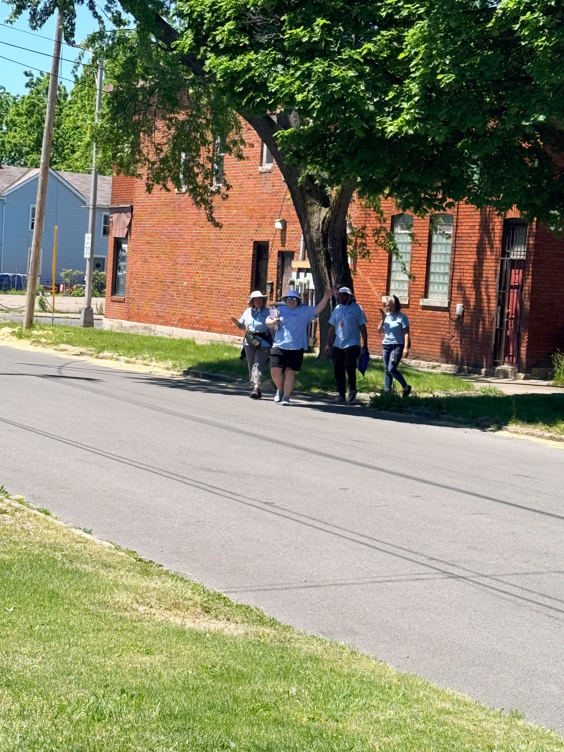 Four people stand on a sidewalk beside a red brick building under a tree on a sunny street.