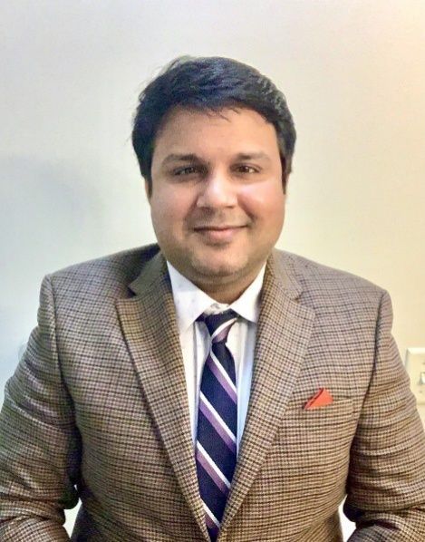 Portrait of a smiling person in a gray plaid suit, striped tie, and red pocket square against a plain background