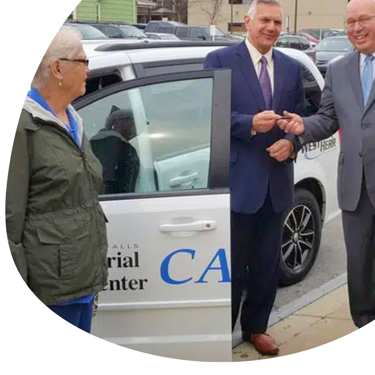 People standing by an open silver car in a parking lot, with two men in suits talking.