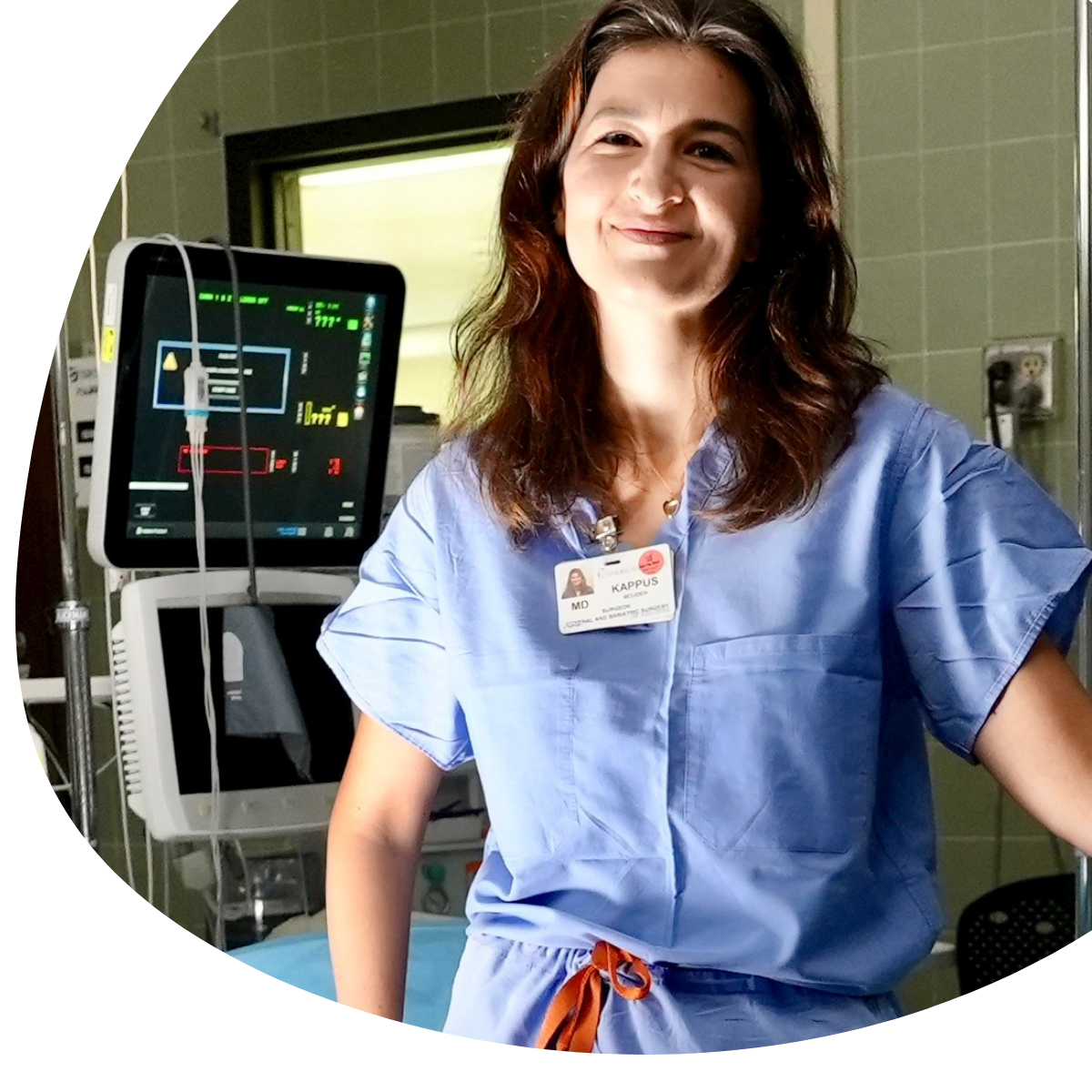 Hospital staff member in blue scrubs smiling beside a patient monitor in a clinical room
