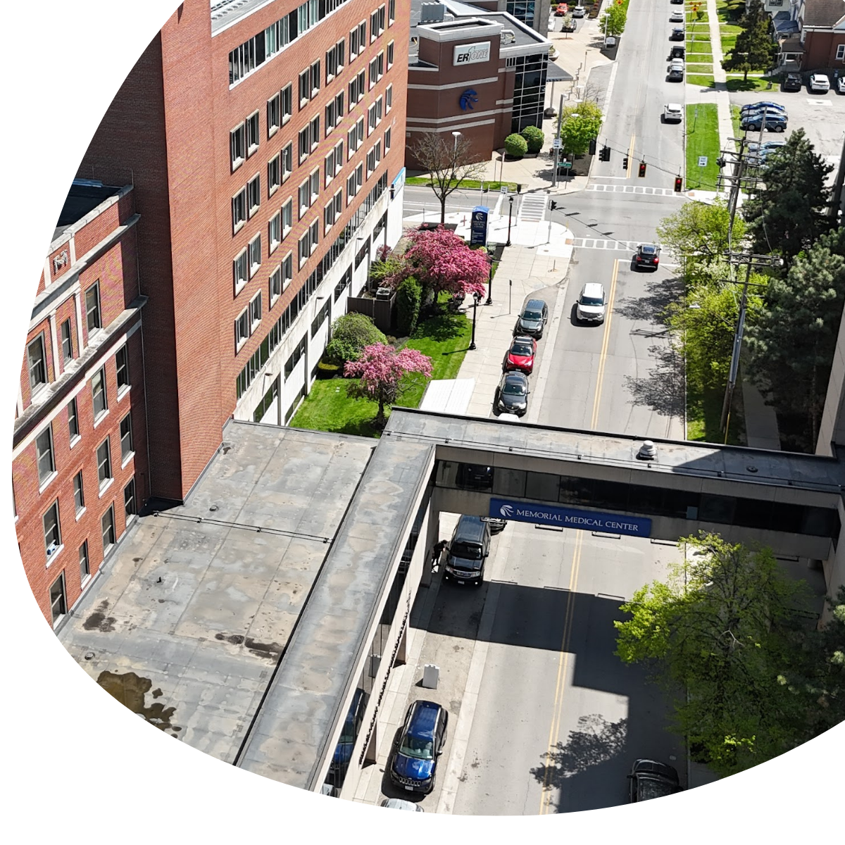 Aerial view of a city street beside brick buildings, with cars, trees, and a skybridge over the road.