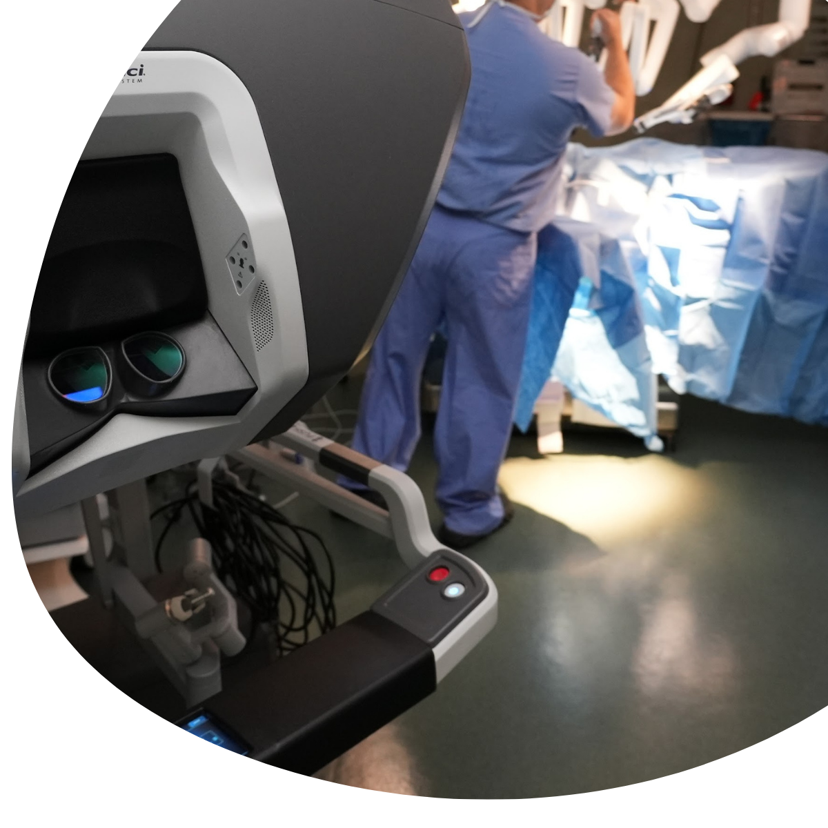 Robotic surgical console in foreground, with surgeons in blue scrubs working in the operating room.