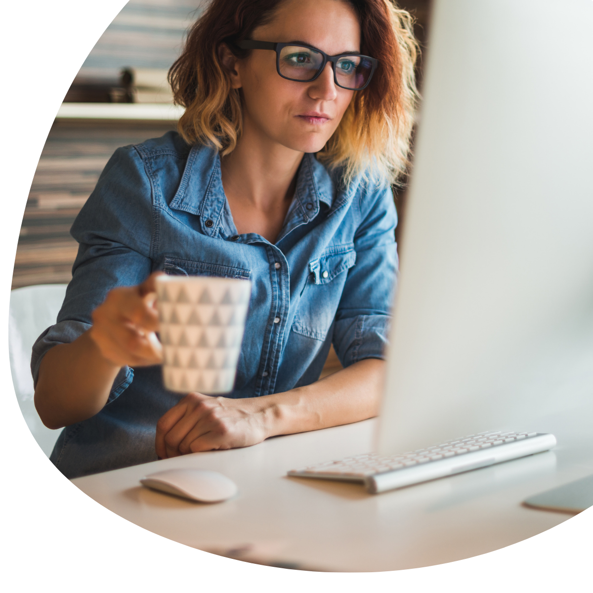 Woman in denim shirt and glasses working at a laptop, holding a mug at a desk