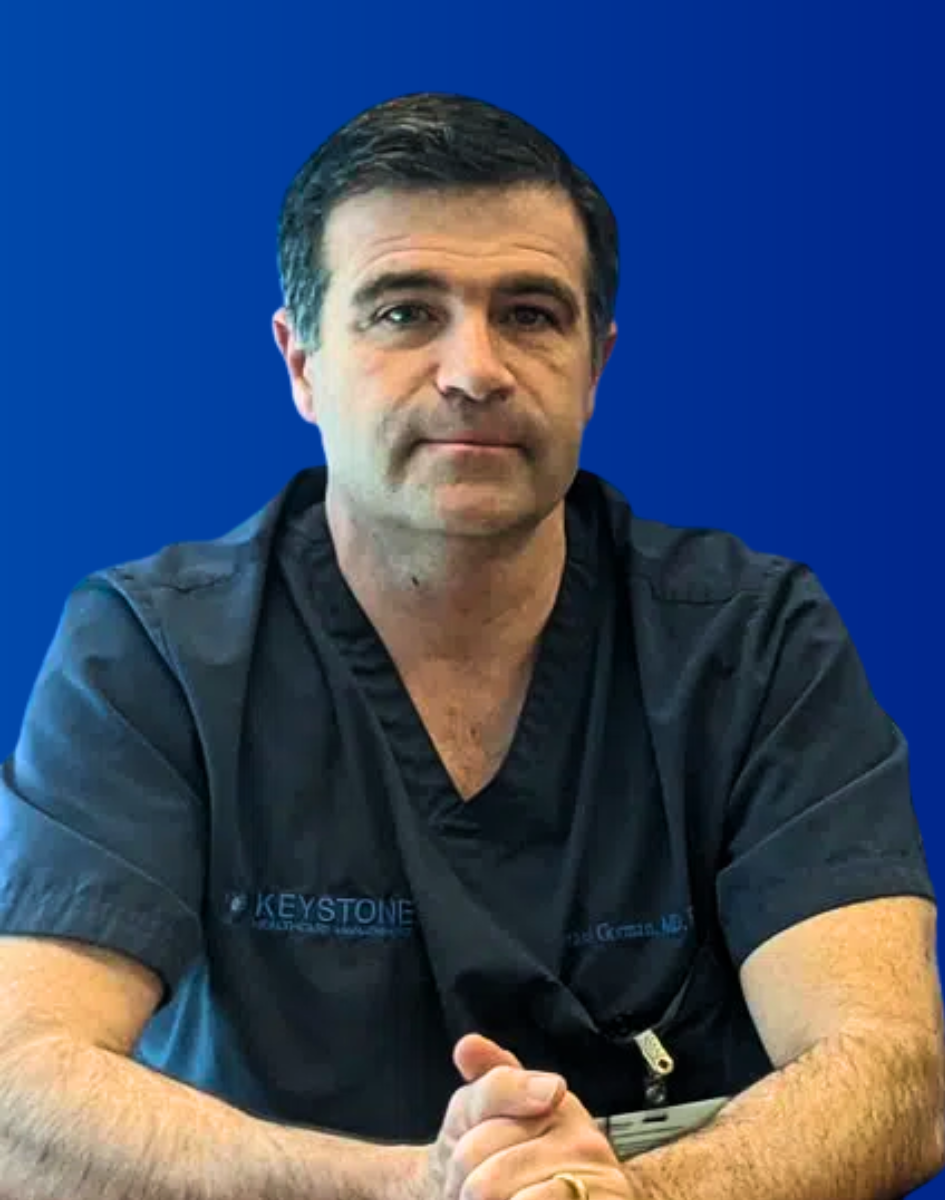 Man in dark scrubs seated against a blue background, facing forward with hands clasped.