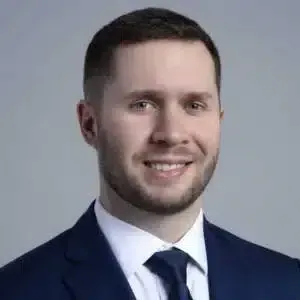 Studio headshot of a smiling man in a navy suit and tie on a gray background