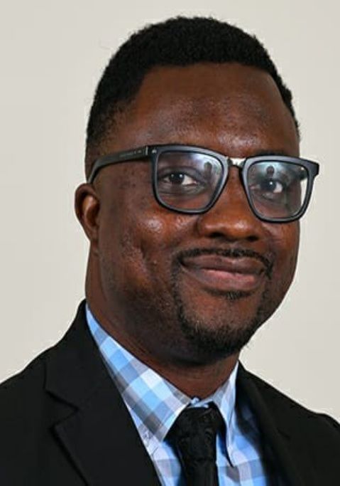 Portrait of a man in glasses wearing a black suit, blue plaid shirt, and black tie against a light background
