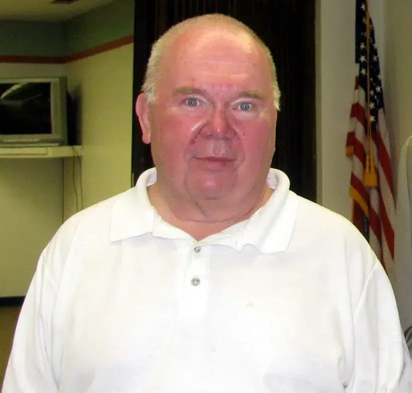 Man in a white polo shirt standing indoors beside an American flag