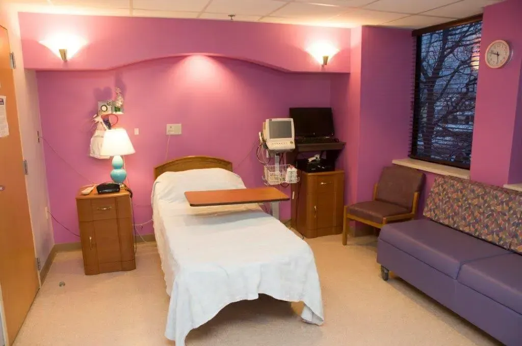 Pink hospital room with a bed, bedside lamp, computer monitor, chair, and sofa